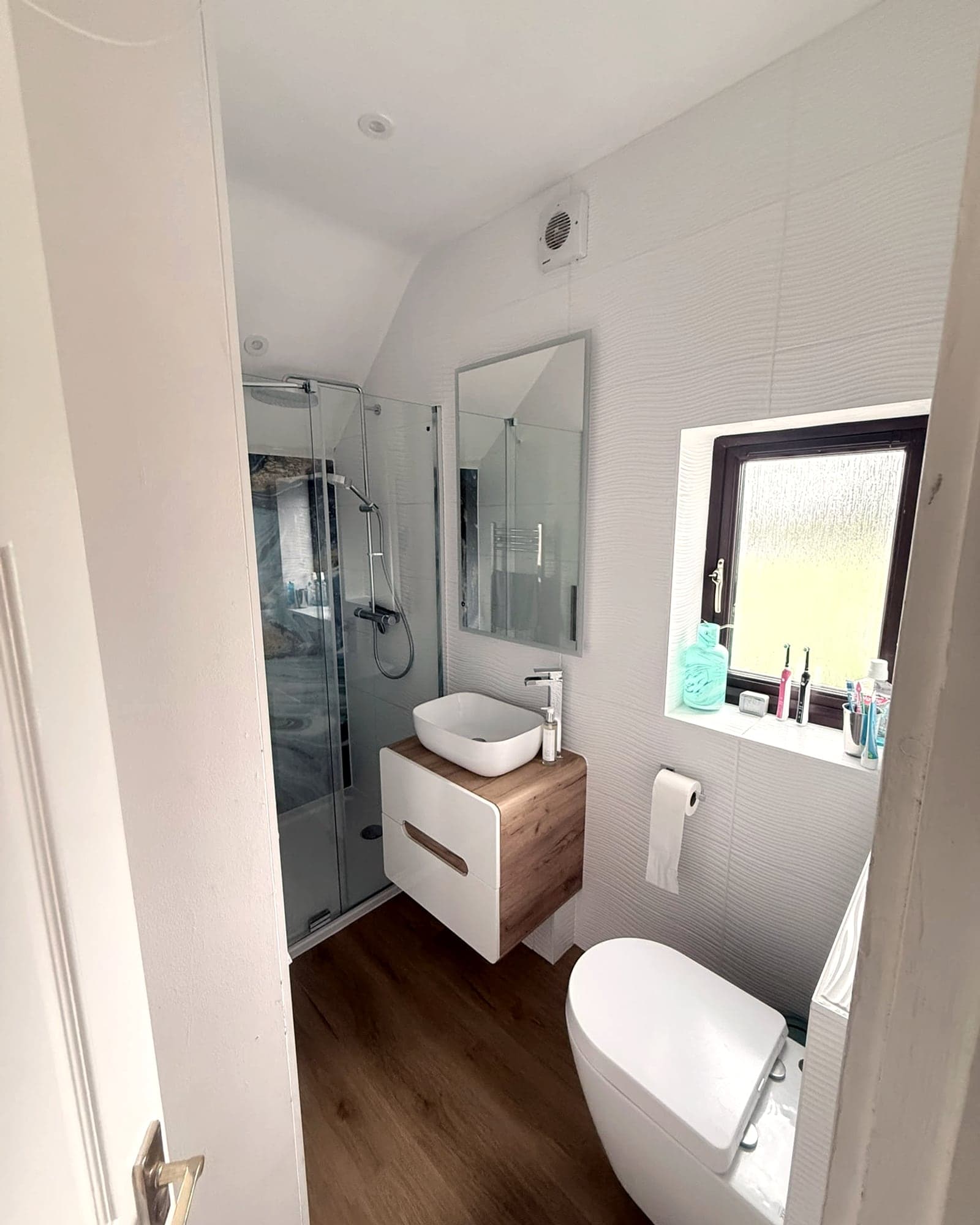 A small en suite: a glass shower, a vessel basin on a wood-effect unit, and a window